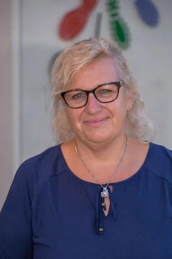 Eine Frau mit blonden Haaren, die eine Brille und ein blaues Hemd trägt und in die Kamera lächelt.