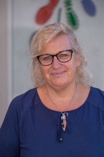 Eine Frau mit blonden Haaren, die eine Brille und ein blaues Hemd trägt und in die Kamera lächelt.