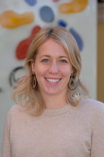 Eine Frau mit blonden Haaren, die ein beiges Sweatshirt trägt, lächelt fröhlich mit den Zähnen. Sie trägt goldene Ohrringe mit Perlen.