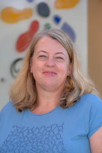 Eine Frau mit blonden Haaren und einem blauen Shirt lächelt und schaut in die Kamera.