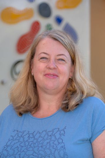 Eine Frau mit blonden Haaren und einem blauen Shirt lächelt und schaut in die Kamera.