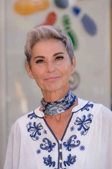 Eine Frau mit kurzen grauen Haaren und einem blauen Schal lächelt in die Kamera. Sie trägt ein weißes Oberteil mit blauer Blumenstickerei.
