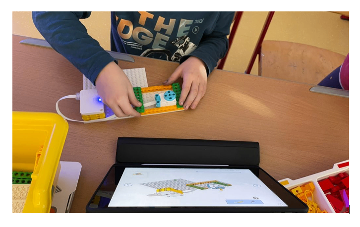 Ein Kind benutzt ein Tablet, um eine LEGO-Struktur zu bauen, mit einem angeschlossenen Gerät auf dem Tisch.