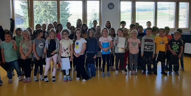 Eine Gruppe von Kindern, in Freizeitkleidung gekleidet, steht in einer Reihe in einem Klassenzimmer, hält Urkunden und posiert für ein Foto.