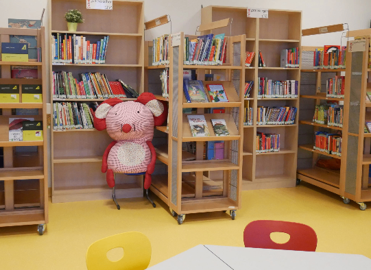 Eine Bibliothek mit Holzregalen voller Bücher. Ein rosa Plüschtier sitzt auf einem Stuhl in der Mitte. Der Boden ist gelb.
