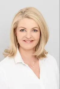 Eine Frau mit kurzen blonden Haaren und einem weißen Blusenhemd, lächelt und posiert für ein Foto.