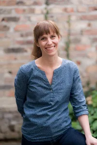 Eine Frau mit kurzen Haaren und blauen Augen lächelt und steht vor einer Ziegelmauer, in einem blauen gepunkteten Oberteil.