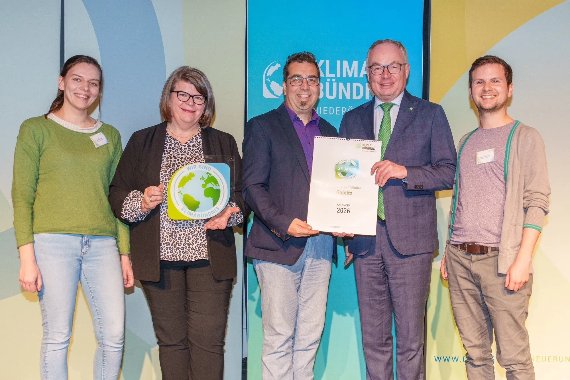 Fünf Personen stehen nebeneinander, lächeln und halten ein Zertifikat. Die beiden Frauen auf der linken Seite halten eine Plakette mit einem Globus darauf. Der Mann auf der rechten Seite hält ein Zertifikat. Der Hintergrund ist eine mehrfarbige Kulisse.