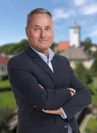 Ein Mann in einem Anzug steht draußen mit verschränkten Armen und lächelt. Er trägt eine Uhr. Im Hintergrund befindet sich eine kleine Stadt mit einem Turm.