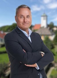 Ein Mann in einem Anzug steht draußen mit verschränkten Armen und lächelt. Er trägt eine Uhr. Im Hintergrund befindet sich eine kleine Stadt mit einem Turm.