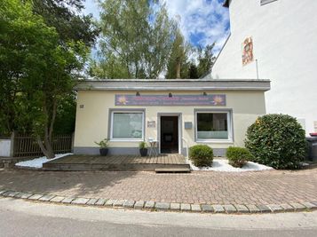 Ein kleines Gebäude mit einer Holzveranda und einem Schild, das 'Fukusase-Gabultz Jennifer Steiner' steht. Topfpflanzen sind auf der Veranda.