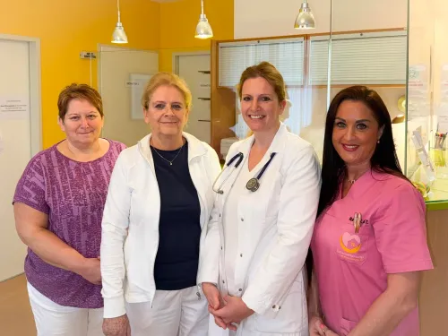 Vier Frauen stehen lächelnd in einem Raum zusammen und posieren für ein Foto. Zwei tragen weiße Laborkittel und Stethoskope, eine trägt ein rosa Hemd und eine trägt ein lila Oberteil.