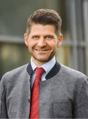 Ein Mann mit grauem Pullover, roter Krawatte und weißem Hemd lächelt und scheint für ein Foto zu posieren.