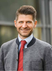 Ein Mann mit grauem Pullover, roter Krawatte und weißem Hemd lächelt und scheint für ein Foto zu posieren.