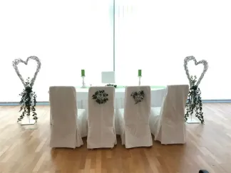 Ein runder weißer Tisch ist von Stühlen umgeben, die mit weißem Stoff und einer Girlande auf dem Rücken bedeckt sind.