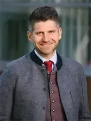Ein lächelnder Mann in einem grauen Mantel mit schwarzem Kragen und einer roten Krawatte mit Knöpfen.