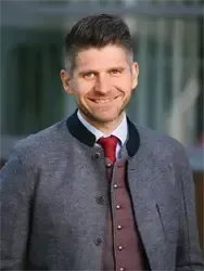 Ein lächelnder Mann in einem grauen Mantel mit schwarzem Kragen und einer roten Krawatte mit Knöpfen.
