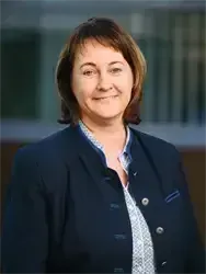 Eine Frau in einem dunkelblauen Blazer und einem weißen gemusterten Hemd lächelt und posiert für ein Foto.