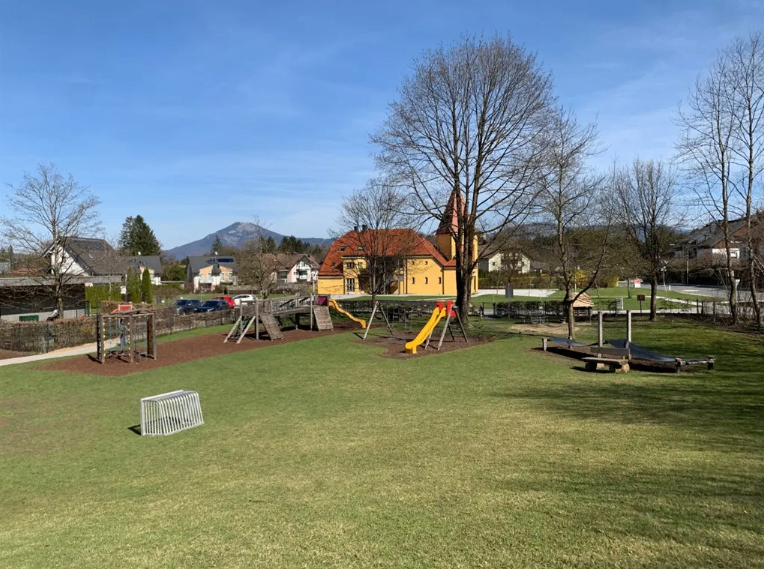 Ein Spielplatz im Freien mit einem Rutschvergnügen und einem Klettergerüst auf einem grünen Rasen. Im Hintergrund befinden sich Häuser, eine Kirche und eine Bergkette.