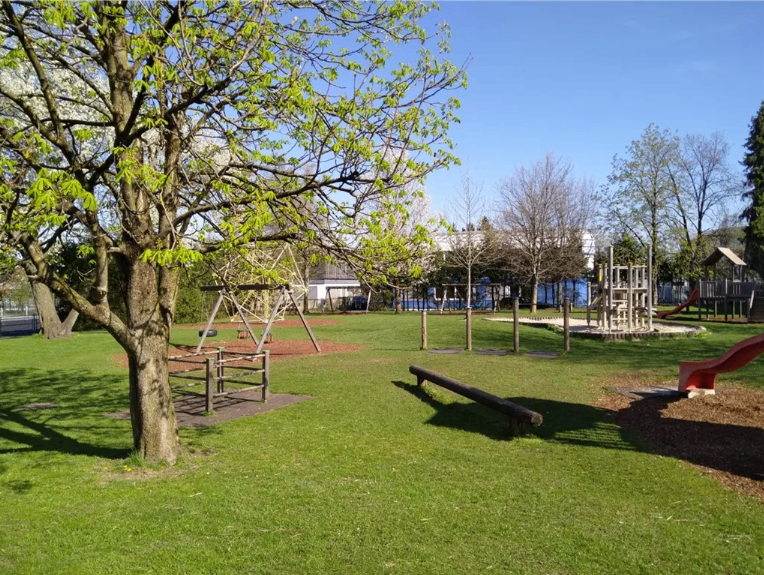 Ein Park mit einem Spielplatz. Es gibt Rutschen, Schaukeln und eine Holzbank. Der Rasen ist grün und die Bäume haben Blätter.
