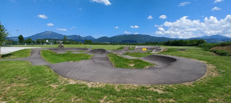 Luftaufnahme eines Skateboardparks mit Rampen und Bowls. Im Hintergrund sind Berge und ein bewölkter Himmel zu sehen.