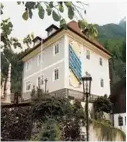Ein großes weißes Gebäude mit einem braunen Dach steht in einer ländlichen Gegend, umgeben von Grün und Bergen.