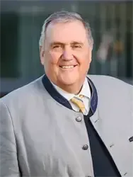 Ein Mann mit grauem Haar trägt einen grauen Mantel und eine gelbe Krawatte und lächelt in die Kamera.