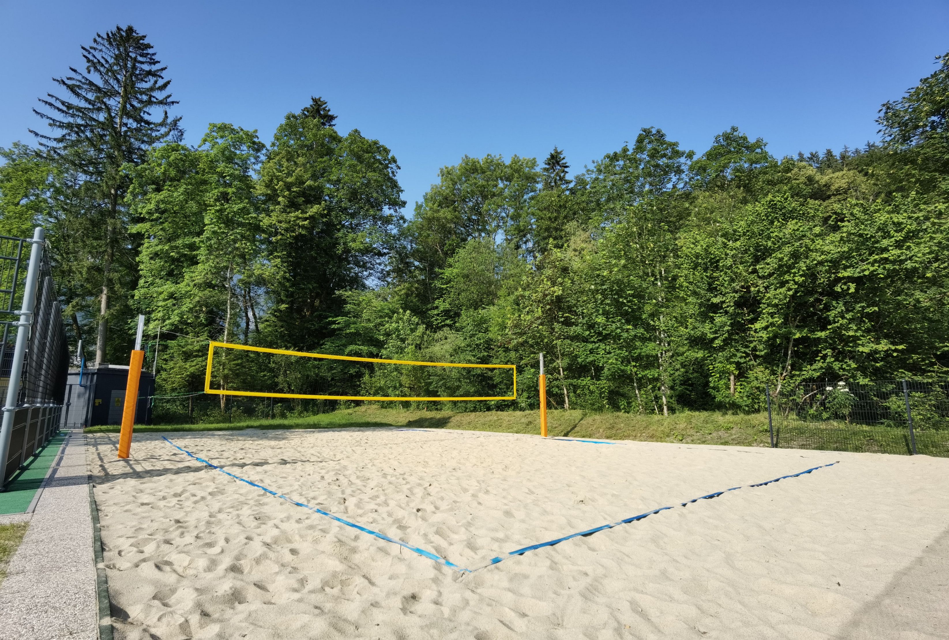 Ein Volleyballfeld steht auf einem sandigen Feld, mit einem gelben Netz, das die beiden Seiten trennt. Das Gebiet ist von üppigem grünem Laub umgeben.