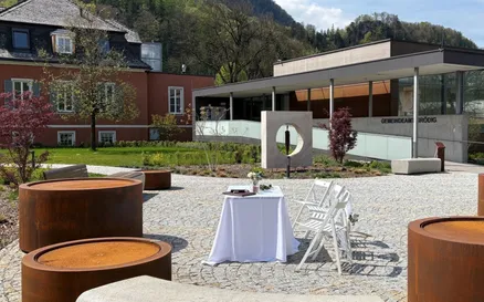 Ein weißer Tisch mit einer weißen Tischdecke ist im Freien mit Stühlen aufgestellt. Ein Gebäude mit einer Glaswand im Hintergrund, mit einem Berg in der Ferne.