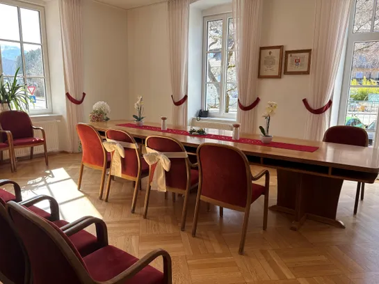 Ein langer Tisch mit sechs Stühlen steht in einem Raum mit weißen Wänden und Holzböden. Jeder Stuhl ist mit einem weißen Band verziert. Auf dem Tisch stehen Blumen in Töpfen und Bilderrahmen an der Wand.