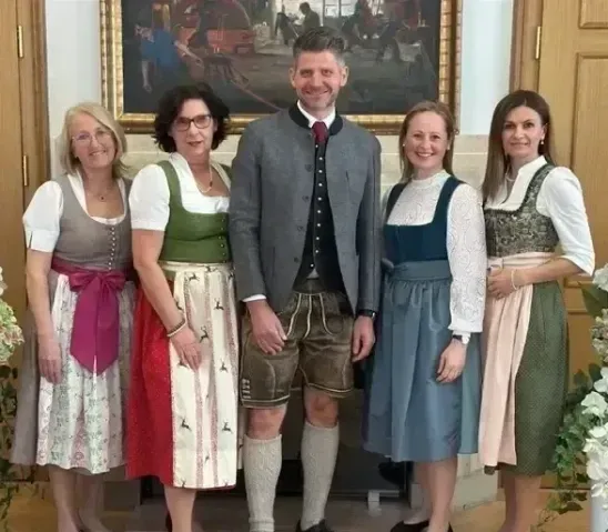 Fünf Personen, drei Frauen und zwei Männer, stehen lächelnd vor einem Gemälde. Die Männer tragen traditionelle bayerische Kleidung.