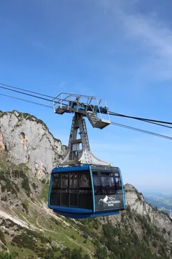 Eine blaue Seilbahn fährt einen steilen Berg hinauf, mit einem klaren blauen Himmel im Hintergrund. Die Seilbahn ist mit Untersberg Bahn beschriftet.