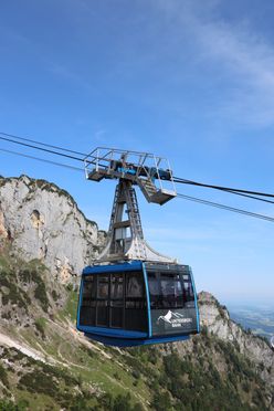 Eine blaue Seilbahn fährt einen steilen Berg hinauf, mit einem klaren blauen Himmel im Hintergrund. Die Seilbahn ist mit Untersberg Bahn beschriftet.