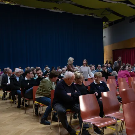 Ein Raum mit einem blauen Vorhang im Hintergrund und einem roten Vorhang an der Seite. Menschen sitzen auf Stühlen, einige stehen. Einige Frauen lächeln, während andere den Blick gesenkt haben.