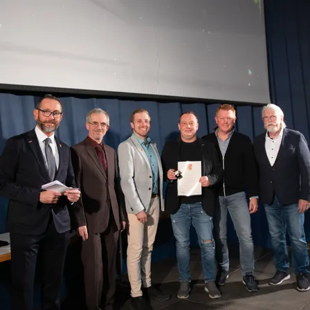 Sechs Männer stehen auf einer Bühne vor einem blauen Vorhang. Ein Mann hält ein Zertifikat. Sie sind alle formell gekleidet.