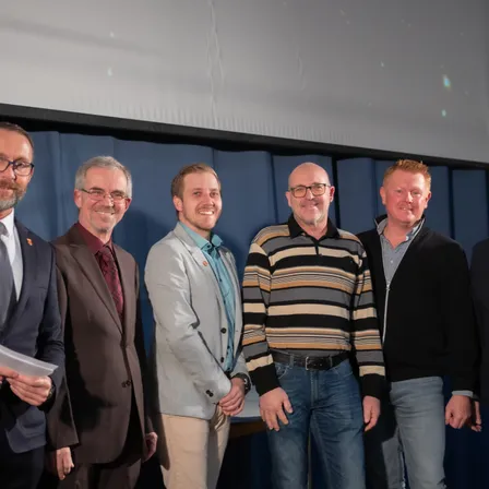 Fünf Männer stehen lächelnd und posieren für ein Foto in einem Konferenzraum. Der Mann auf der linken Seite hält ein Stück Papier.