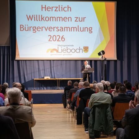 Ein Mann spricht an einem Podium während einer Bürgerversammlung im Jahr 2026. Die Bühne hat eine große Leinwand mit Text, der die Teilnehmer begrüßt.