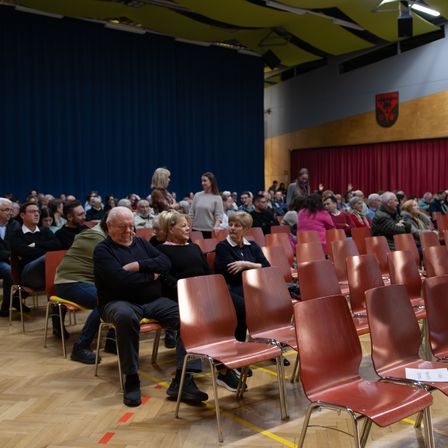 Publikum in Reihen mit einigen Stehplätzen in einem Saal mit Holzböden und roten Stühlen. Eine Frau in Pink steht, und ein blauer Vorhang ist im Hintergrund.