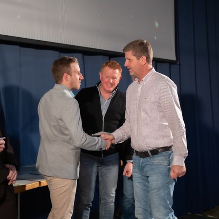 Drei Männer stehen in einem Raum. Zwei schütteln Hände, und einer lächelt und schaut den Mann an, der Hände schüttelt. Hinter ihnen ist eine blaue Vorhang, und eine Person steht auf der linken Seite.