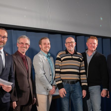 Fünf Männer stehen lächelnd und posieren für ein Foto in einem Konferenzraum. Der Mann auf der linken Seite hält ein Stück Papier.