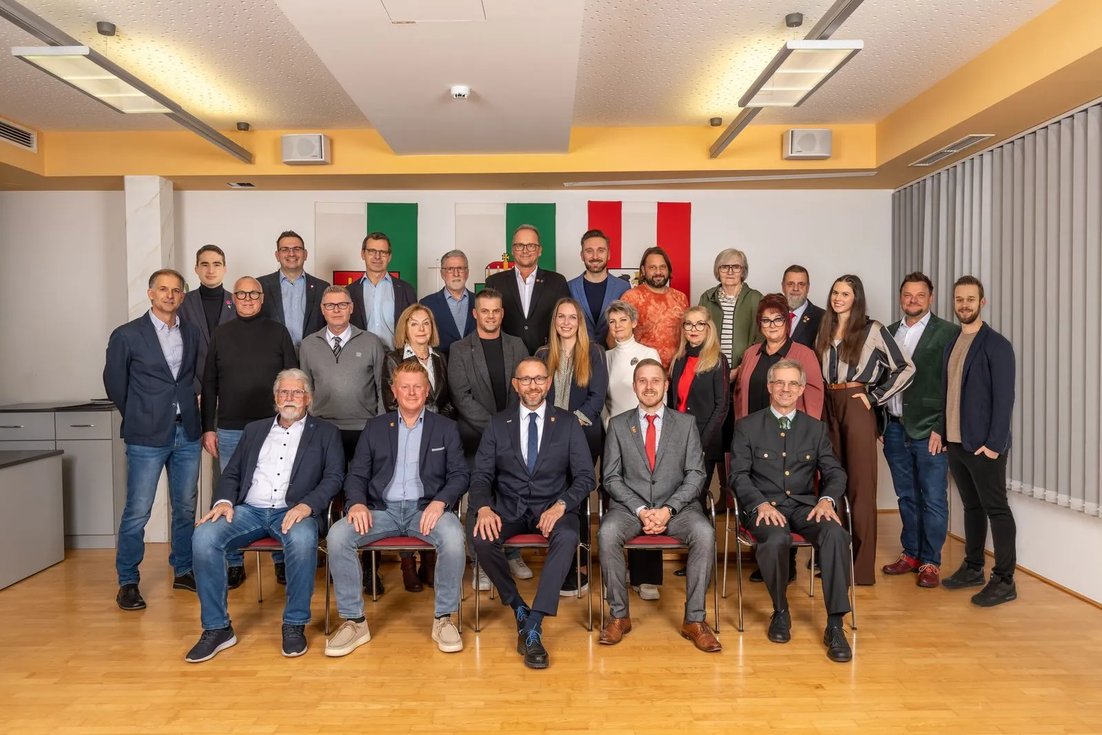 Eine Gruppe von Menschen, darunter Männer und Frauen, posiert für ein Foto in einem Raum mit Holzboden. Sie tragen alle formelle Kleidung. Dahinter befinden sich Fahnen in verschiedenen Farben.