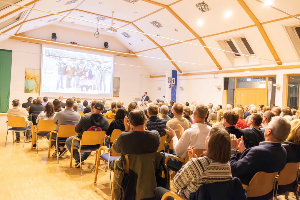 Abschnittsbild: Ein Konferenzsaal mit Holzboden, gefüllt mit Zuhörern, die auf Stühlen sitzen. Ein Mann steht vor einer großen Projektionsleinwand und präsentiert. Der Raum hat Holzbalken und weiße Wände.