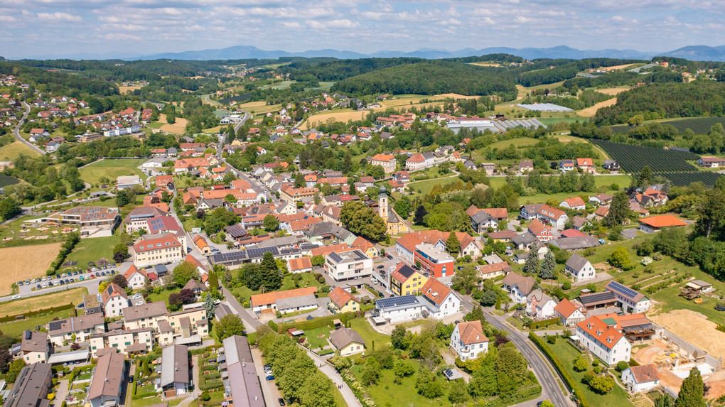  Abschnitt Bild: Luftaufnahme einer kleinen Stadt umgeben von üppigem Grün, Bergen in der Ferne und einer Mischung aus Gebäuden, darunter einige mit Solarmodulen.