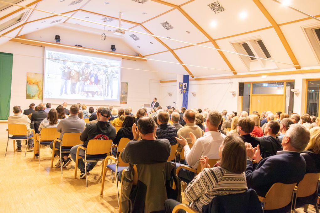  Abschnitt Bild: Ein Konferenzsaal mit Holzboden, gefüllt mit Zuhörern, die auf Stühlen sitzen. Ein Mann steht vor einer großen Projektionsleinwand und präsentiert. Der Raum hat Holzbalken und weiße Wände.