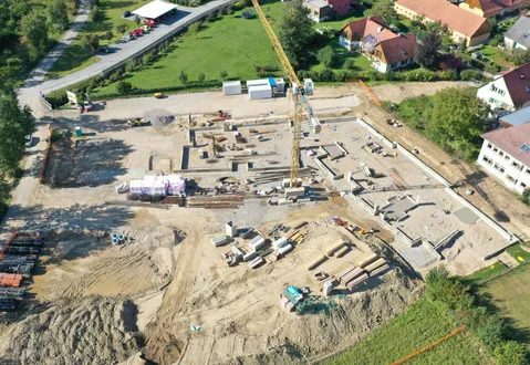 Ein Luftbild eines Baugeländes zeigt verschiedene Gebäude und Strukturen im Bau, wobei ein Kran sichtbar ist. Um das Gelände herum befinden sich Materialhaufen, Maschinen und ein teilweise geräumter Bereich.
