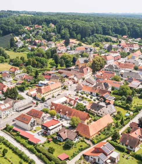 Luftaufnahme eines europäischen Dorfes, umgeben von üppigem Grün und Bäumen. Zahlreiche Häuser mit roten Dächern, einige mit Solarpanels, sind über das Gebiet verteilt. Das Dorf liegt in einem Tal mit Hügeln in der Ferne.