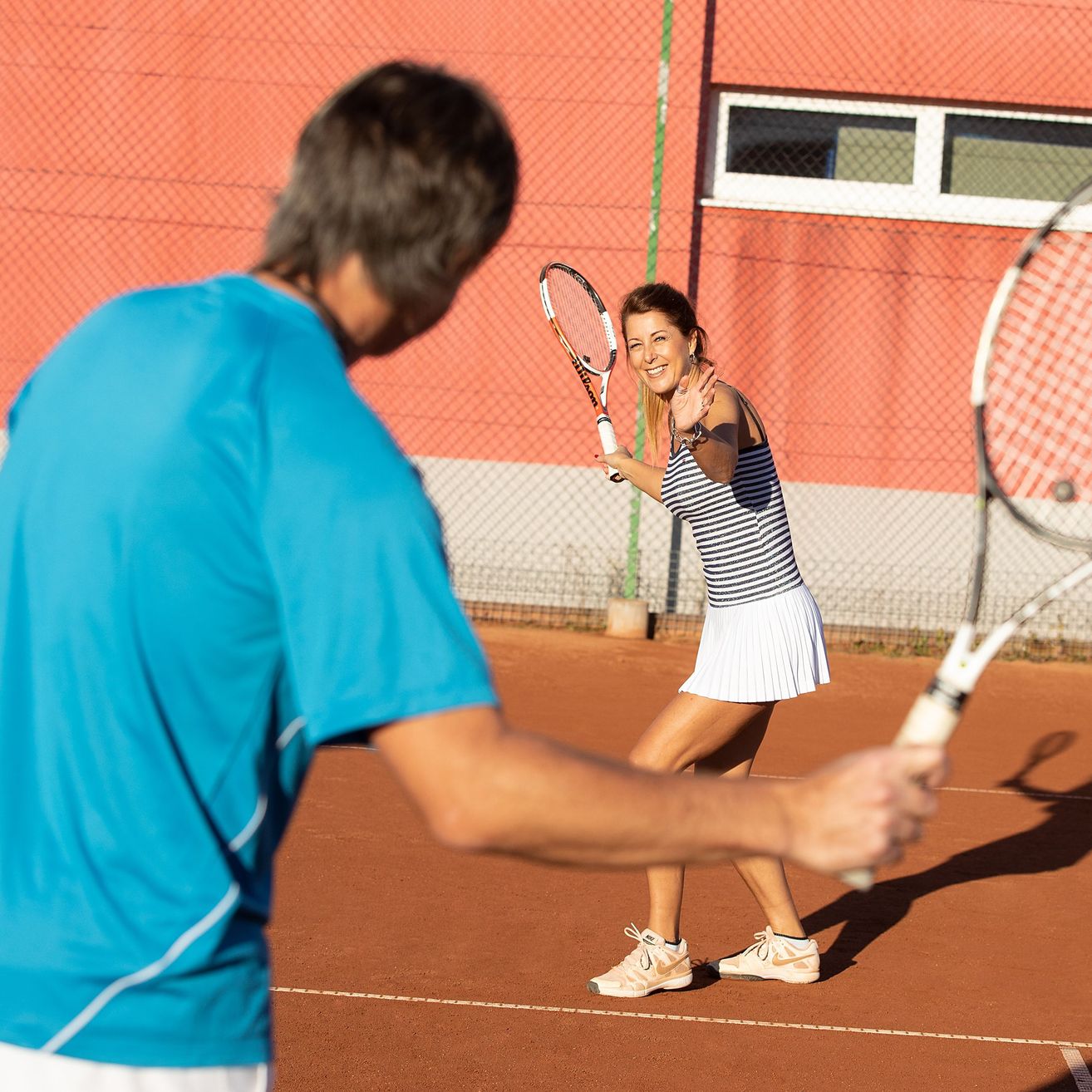 Ein Mann und eine Frau spielen Tennis auf einem Außenplatz, die Frau lächelt und schaut den Mann an.