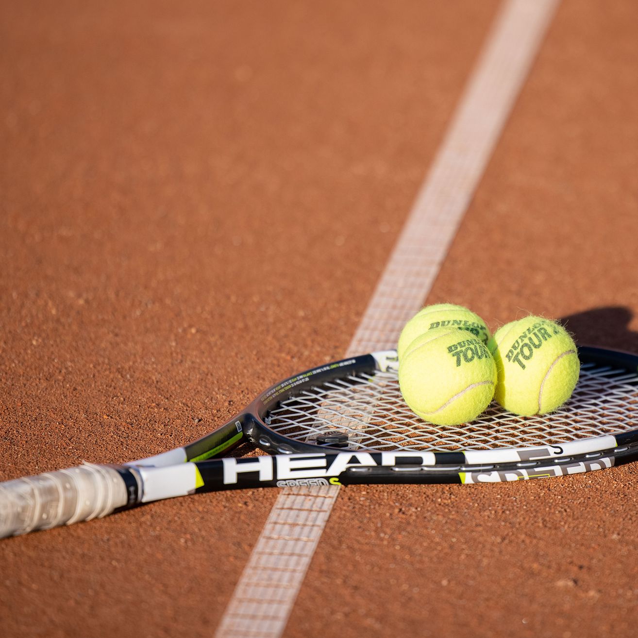 Ein HEAD Tennisschläger liegt auf einem Sandplatz mit zwei Dunlop Tennisbällen.