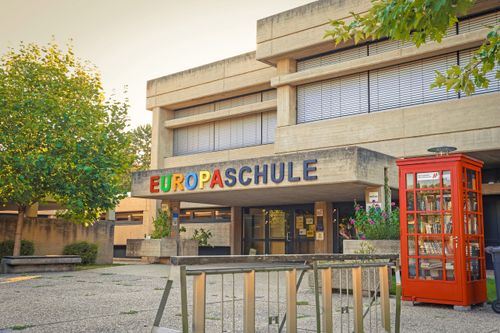Das Äußere des Europaschule-Gebäudes mit moderner Architektur, großen Fenstern und einem bunten Schild über dem Eingang.