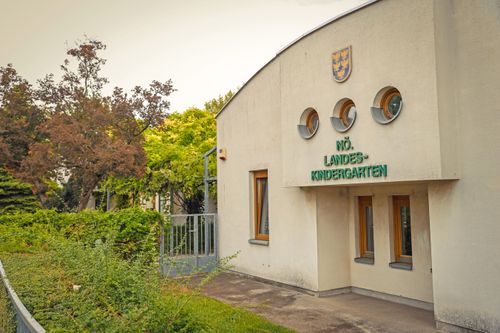 Ein Kindergartengebäude mit einem Schild, das 'Nr. Landes-Kindergarten' über dem Eingang zeigt, mit drei runden Fenstern und einem kleinen Wappen über dem Schild.